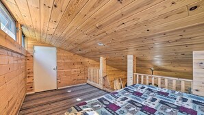 A wooden cabin room with a bed, a door, a window, and a staircase. - Charming Houghton Lake Cottage w/ Backyard! (Prudenville)