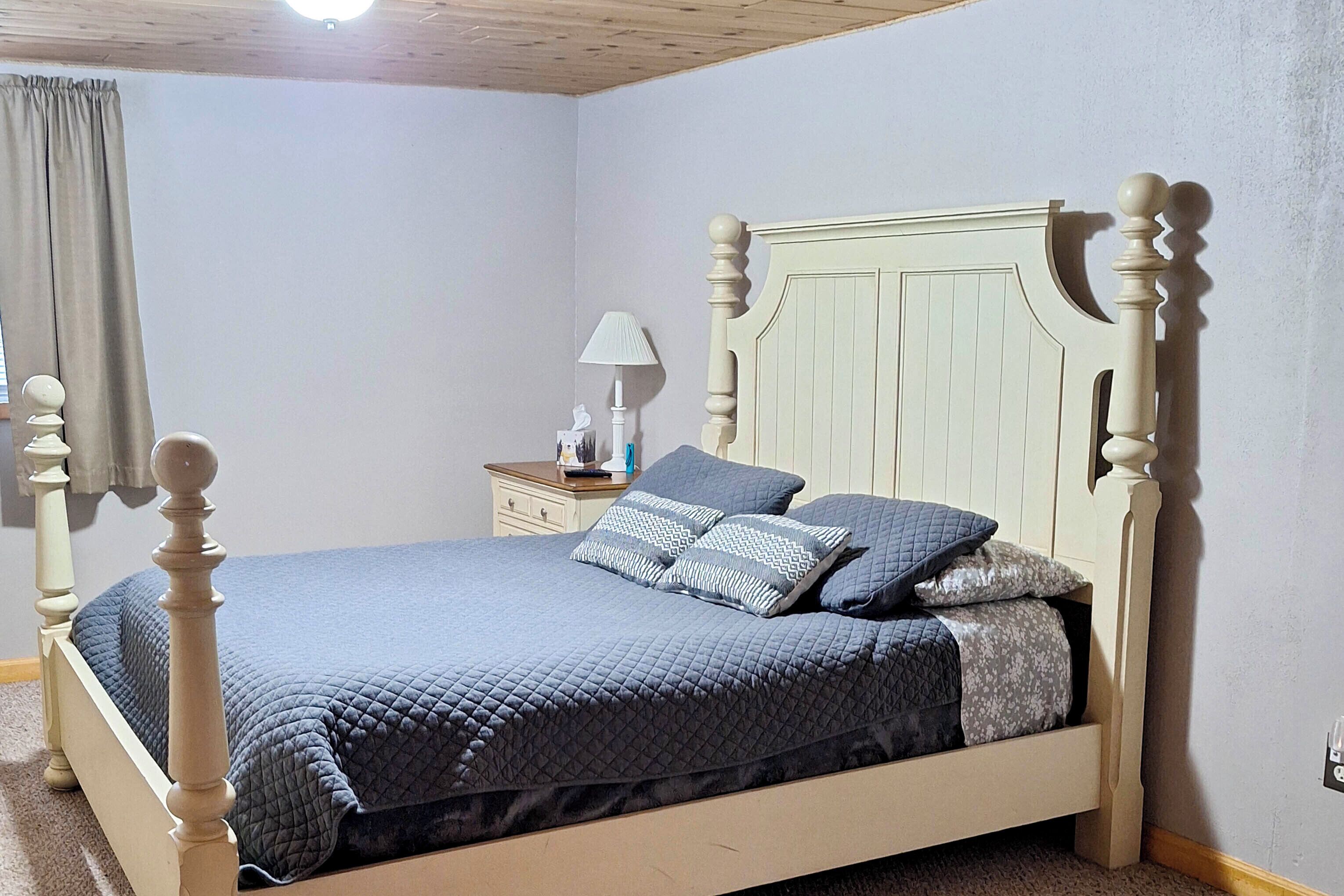 A bedroom with a wooden four-poster bed, a nightstand with a lamp, and a window with curtains.