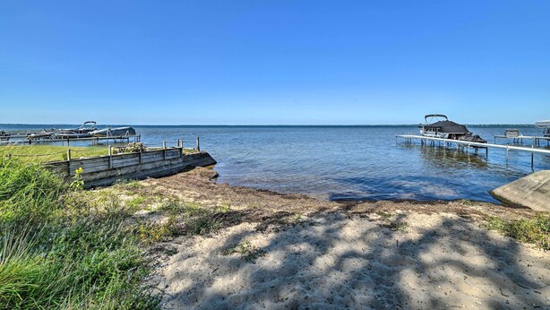 A lakeside view with a dock, a covered area, and a sandy beach. - Charming Houghton Lake Cottage w/ Backyard! (Prudenville)