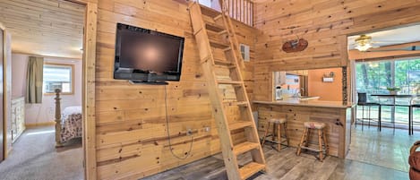 Een houten blokhut van binnen, met een aan de muur bevestigde televisie, een houten trap en een keukenhoek met krukken.