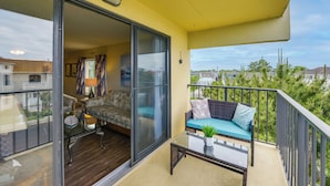 Un balcon avec un canapé et une table en verre, une porte-fenêtre coulissante donnant sur le salon, et une vue sur les maisons et les arbres environnants.