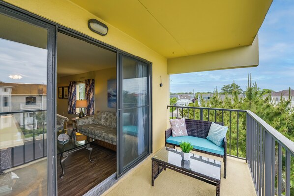 A balcony with a sofa and a glass table, a sliding glass door leading to a living room, and a view of surrounding houses and trees. - Beautiful Brigantine Condo w/ Patio & Beach Access (Brigantine)