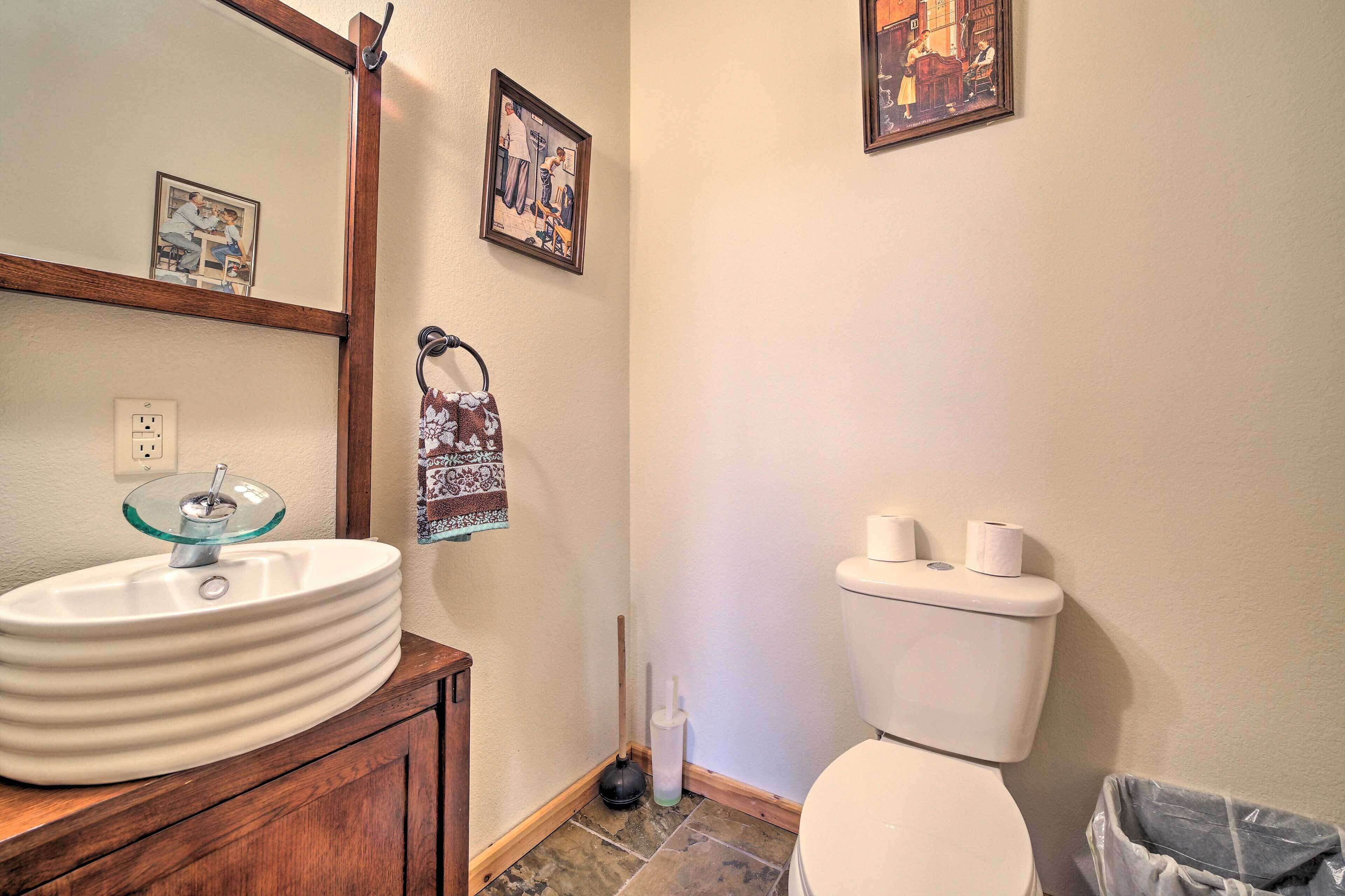 A bathroom with a white sink, toilet, and a mirror. There are framed pictures on the wall and a trash bin.