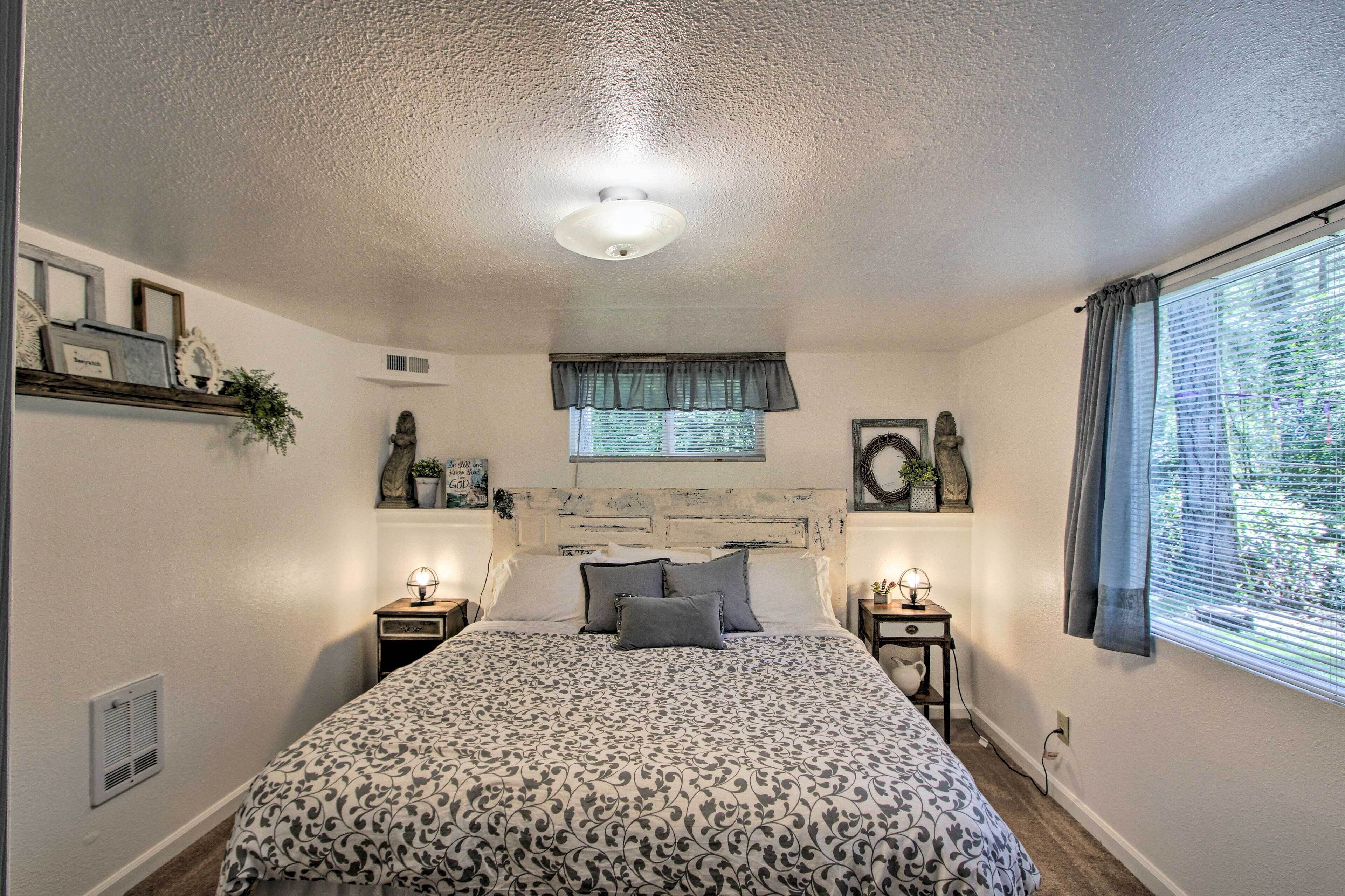 A bedroom with a bed, two bedside tables, a window with curtains, a ceiling fan, and a wall-mounted shelf with decorative items.