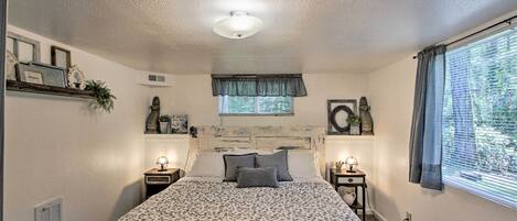 A bedroom with a bed, two bedside tables, a window with curtains, a ceiling fan, and a wall-mounted shelf with decorative items.