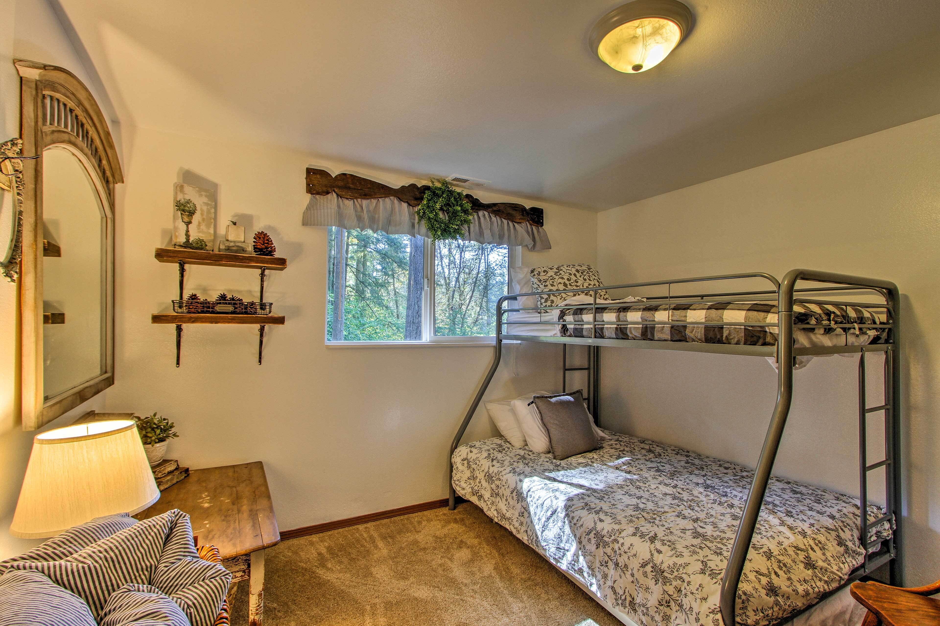 A dormitory room with bunk beds, a mirror, a lamp, a small table, and a window with a view of trees.