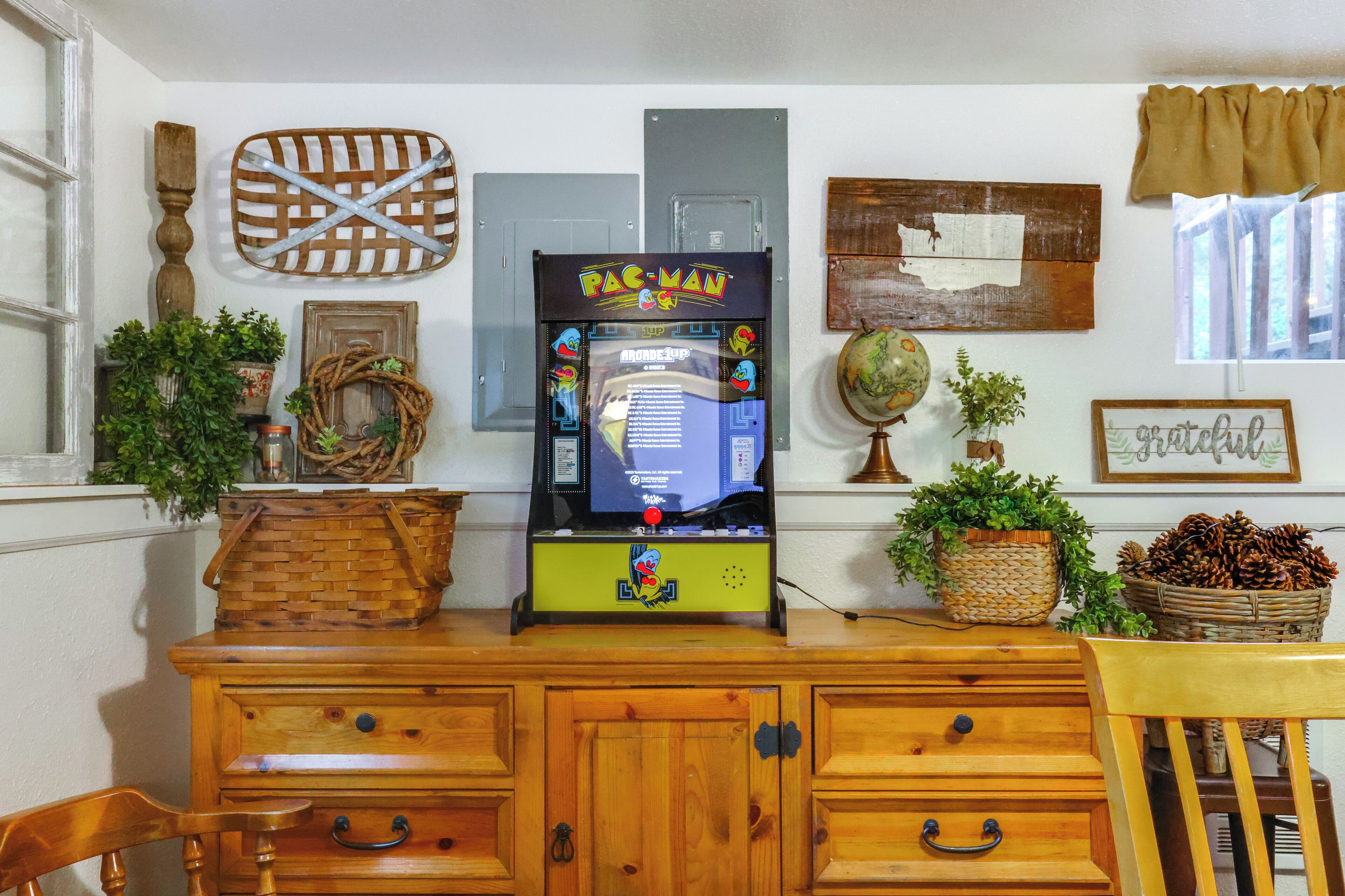 Een kamer met een Pac-Man spelcomputer, houten meubels en diverse decoraties, waaronder een wereldbol, planten en een rieten mand.