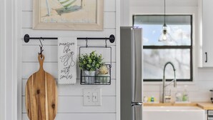 Eine Küche mit einem Edelstahl-Kühlschrank, einem hängenden Schneidebrett aus Holz, einem gerahmten Bild und einem Blumentopf auf einem Metallregal.