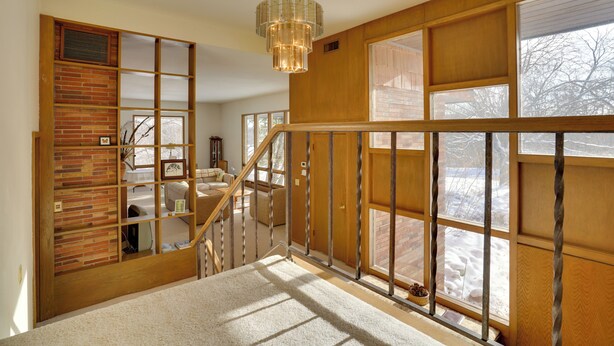 Una escalera con barandillas de madera y un candelabro, que conduce a una habitación con pared de ladrillo y ventanas grandes.