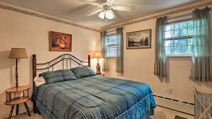 A bedroom with a bed, a ceiling fan, two windows with curtains, a nightstand with a lamp, and a chair. - Saybrook Manor House, Walk to Cove Beach! (Old Saybrook)