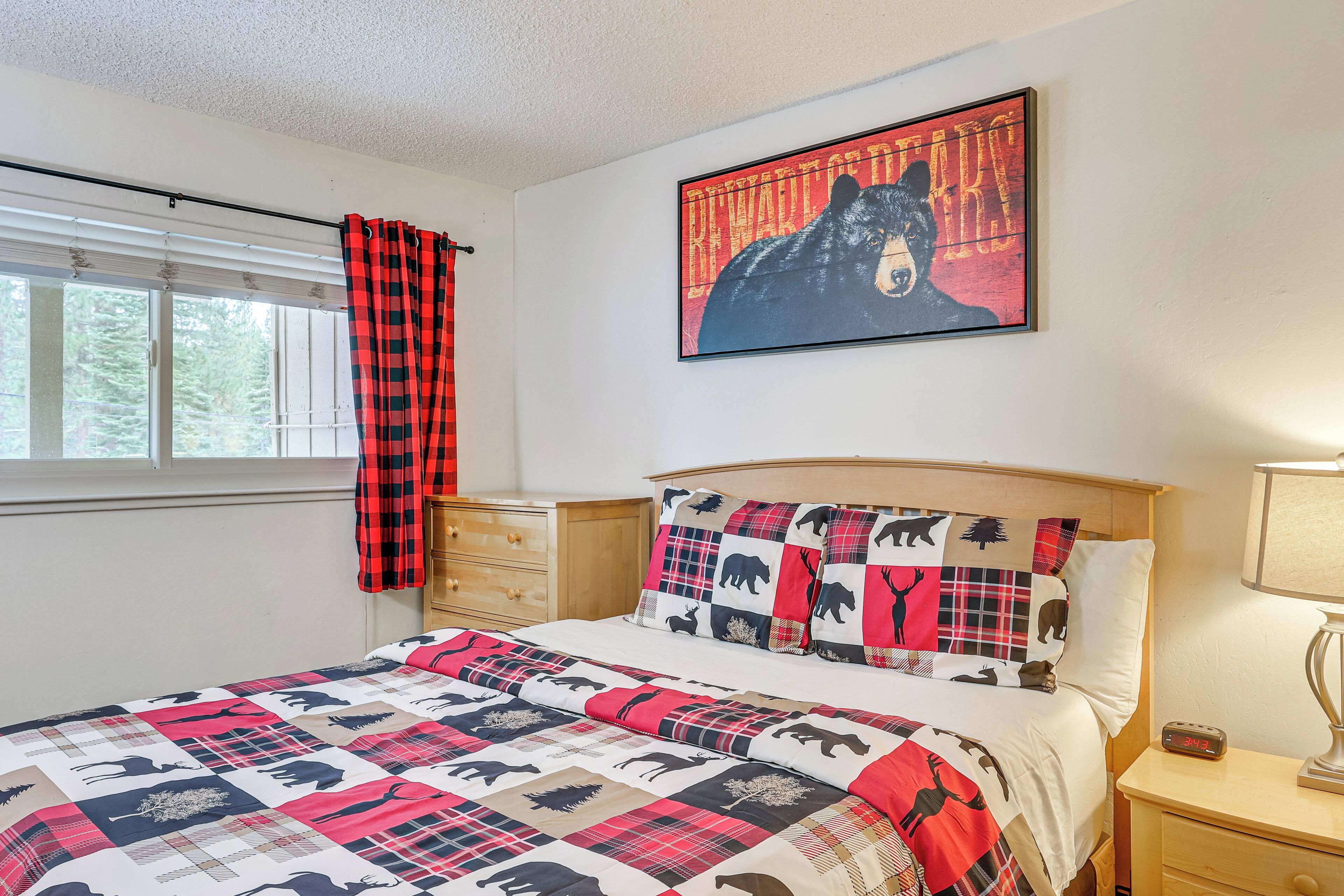 A bedroom with a bed, a nightstand, a lamp, a window with blinds, and a wall with a bear-themed artwork.