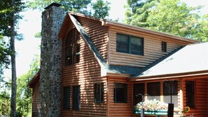 A log cabin with a stone chimney, large windows, and a porch with flowers. - Upscale Fryeburg Cabin: Hot Tub + Billiards Table! (Fryeburg)