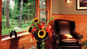 A wooden cabin interior with a large window, a leather chair, a glass table with a vase of flowers, and a framed picture on the wall. - Upscale Fryeburg Cabin: Hot Tub + Billiards Table! (Fryeburg)