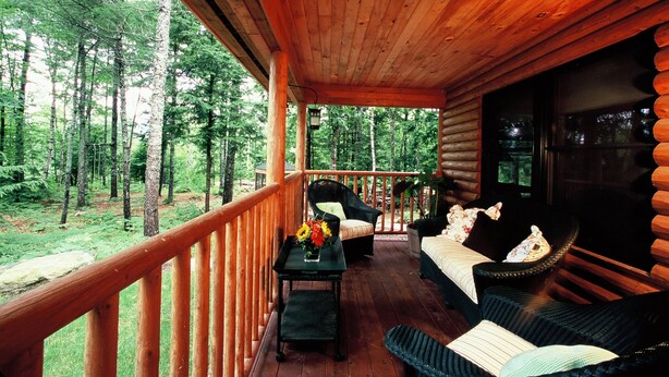 A wooden deck with a view of a forest, a table with a vase of flowers, and two chairs with pillows. - Upscale Fryeburg Cabin: Hot Tub + Billiards Table! (Fryeburg)