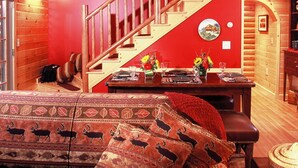 A log cabin interior with a wooden staircase, a dining area with a table set for two, and a cozy seating area with patterned cushions. - Upscale Fryeburg Cabin: Hot Tub + Billiards Table! (Fryeburg)