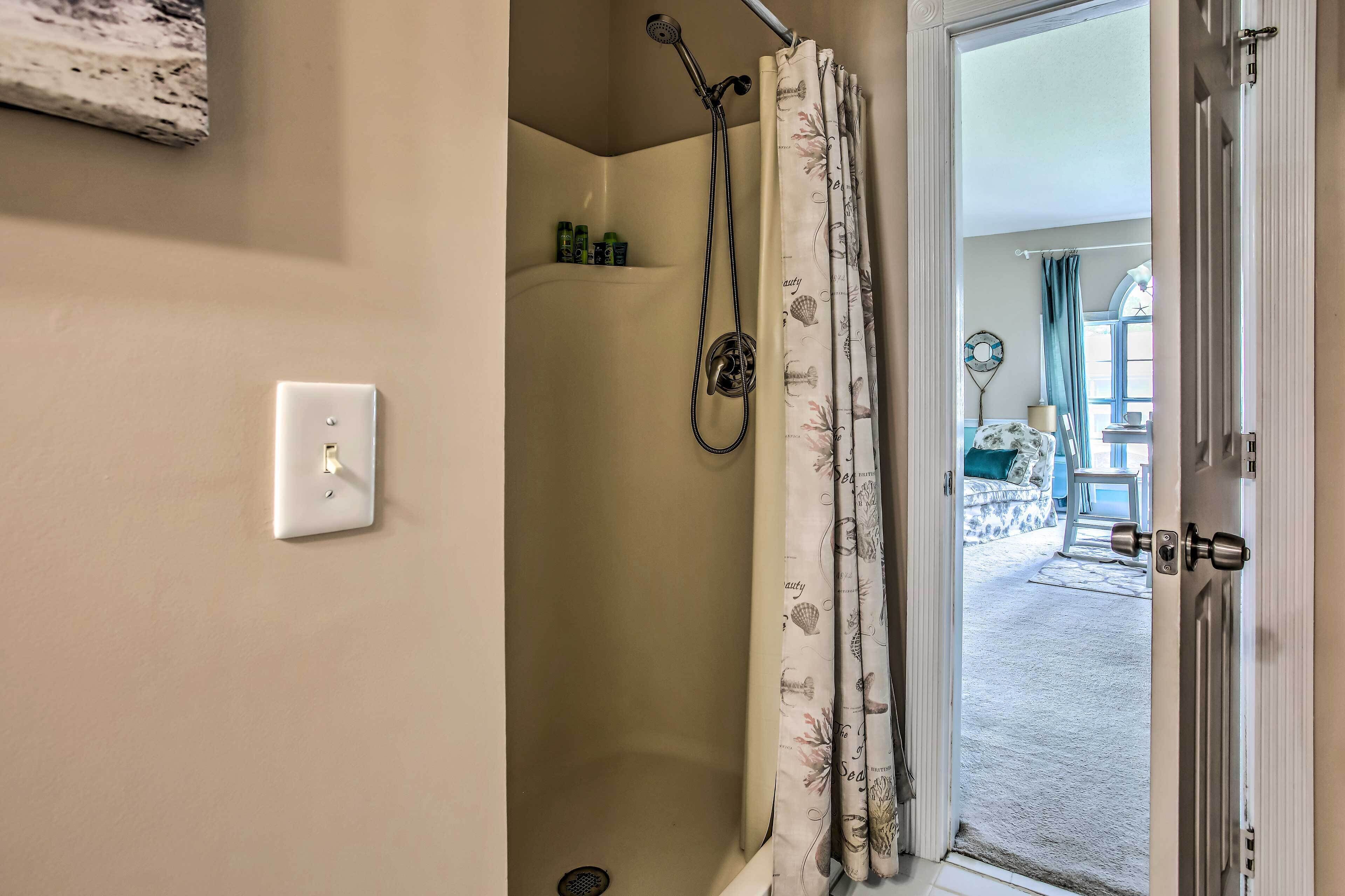 Une salle de bain avec une douche, un mur blanc, une porte et une vue sur une pièce comprenant un lit et une fenêtre.