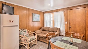 A cozy room with wooden walls, a white refrigerator, a wicker chair, a brown sofa, and a dining table with a placemat. - Cozy Hampton Cottage w/ BBQ - Walk to Ocean! (Hampton)