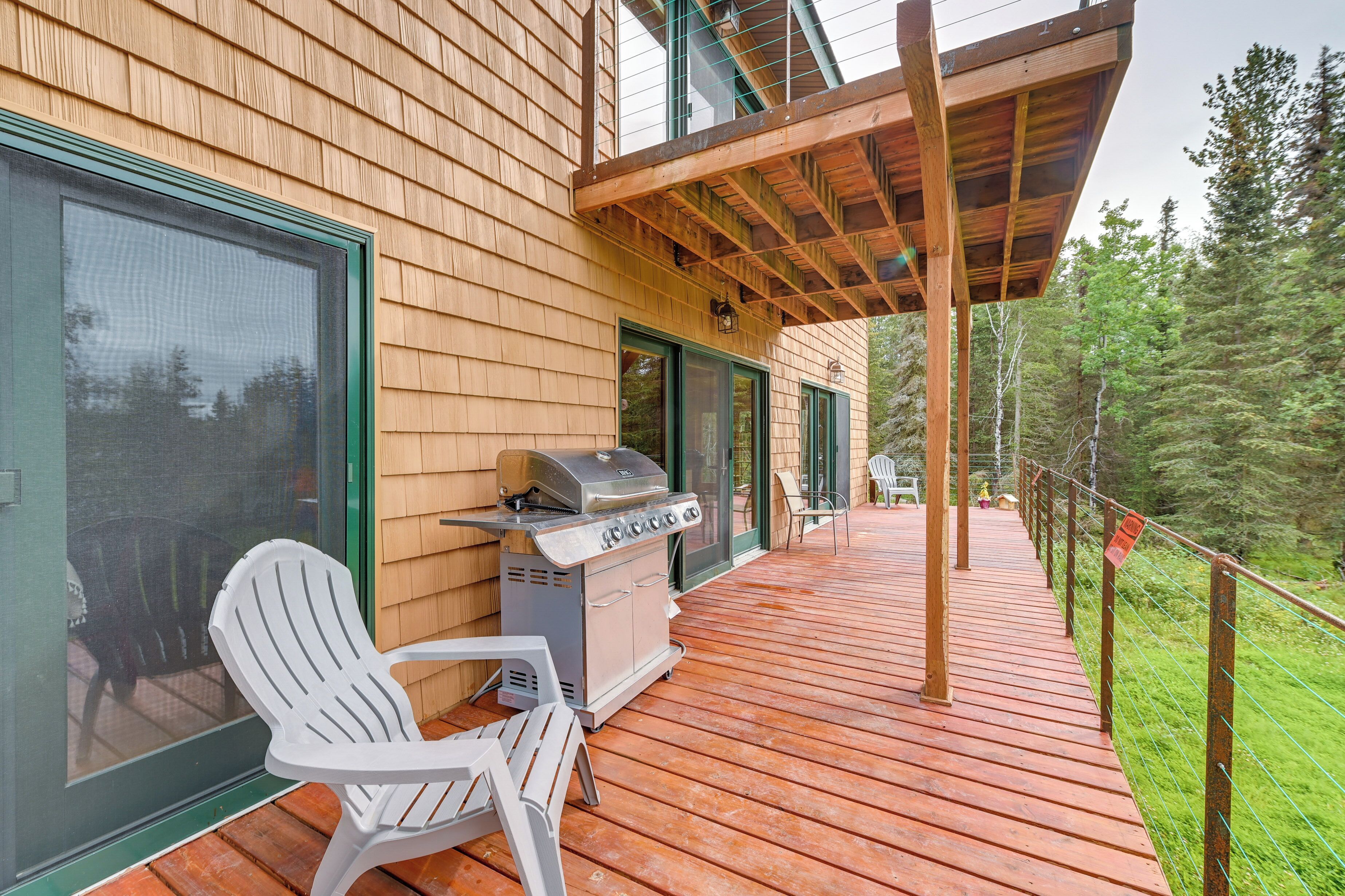 A wooden deck with a grill and white chairs, overlooking a forested area.