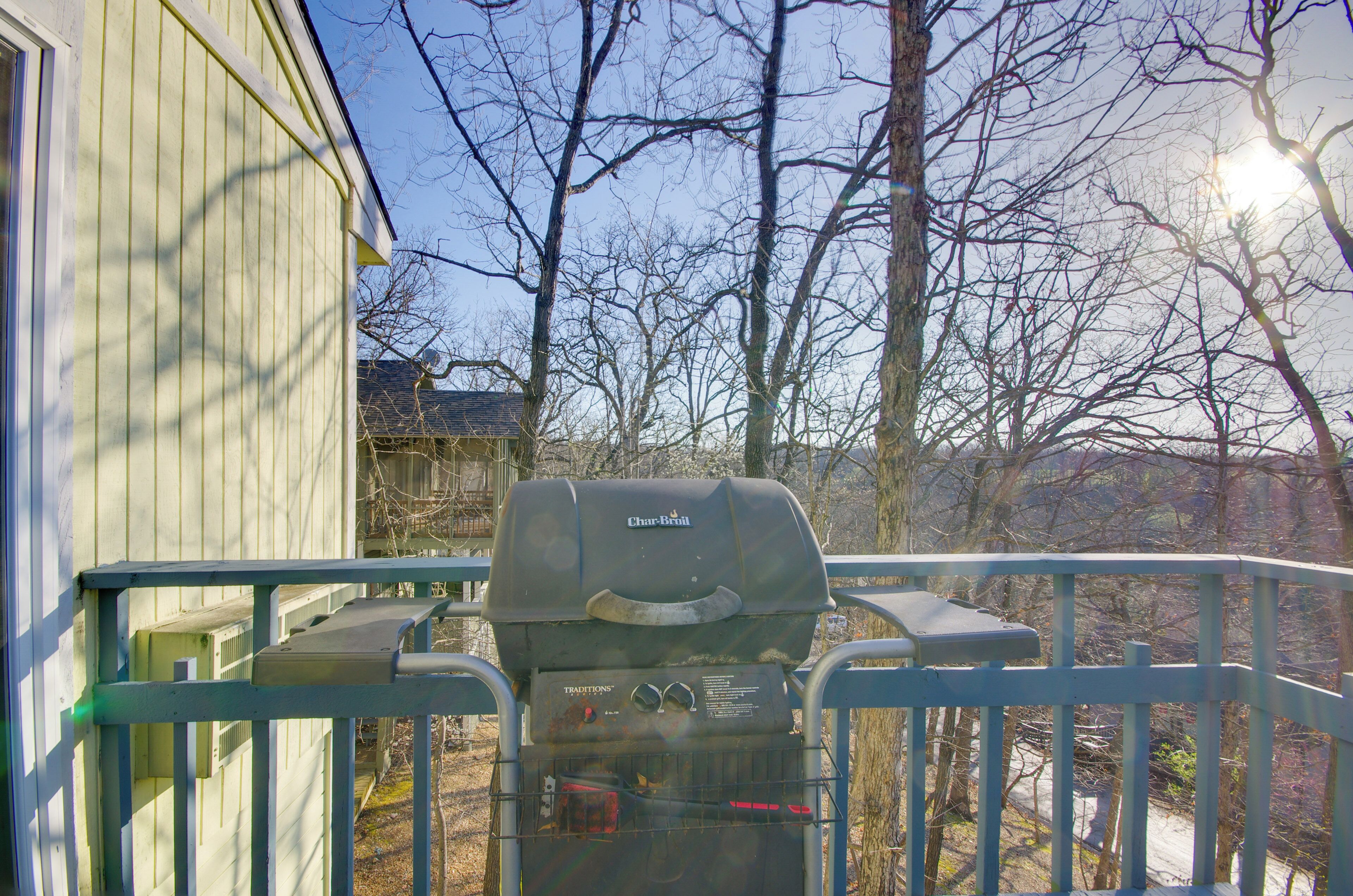 A Char-Bri grill on a balcony with a metal railing, a wooden house in the background, and leafless trees.