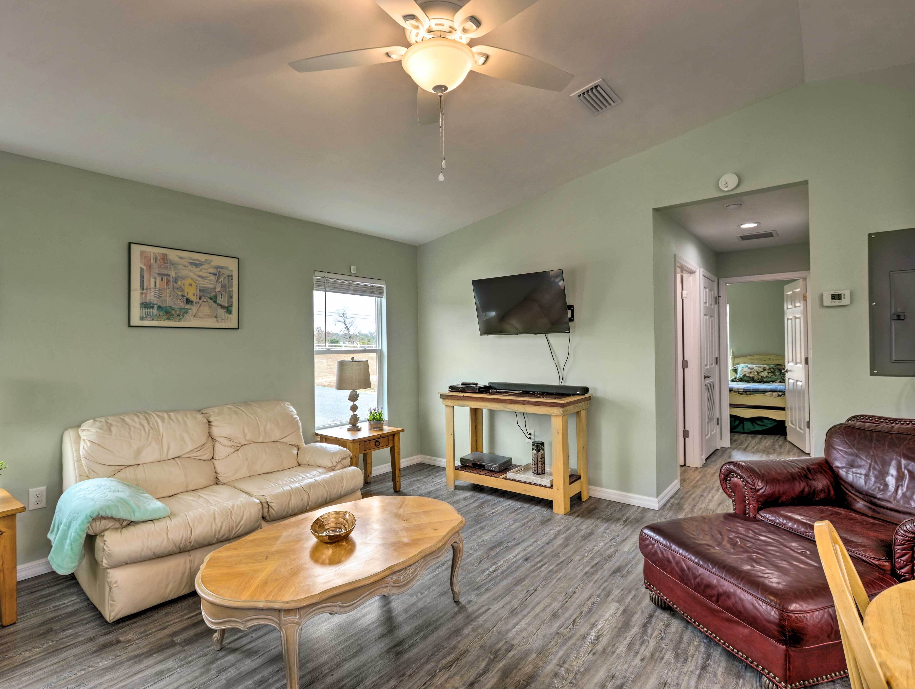 A living room with a beige sofa, a wooden coffee table, a red leather couch, a TV mounted on the wall, and a ceiling fan.