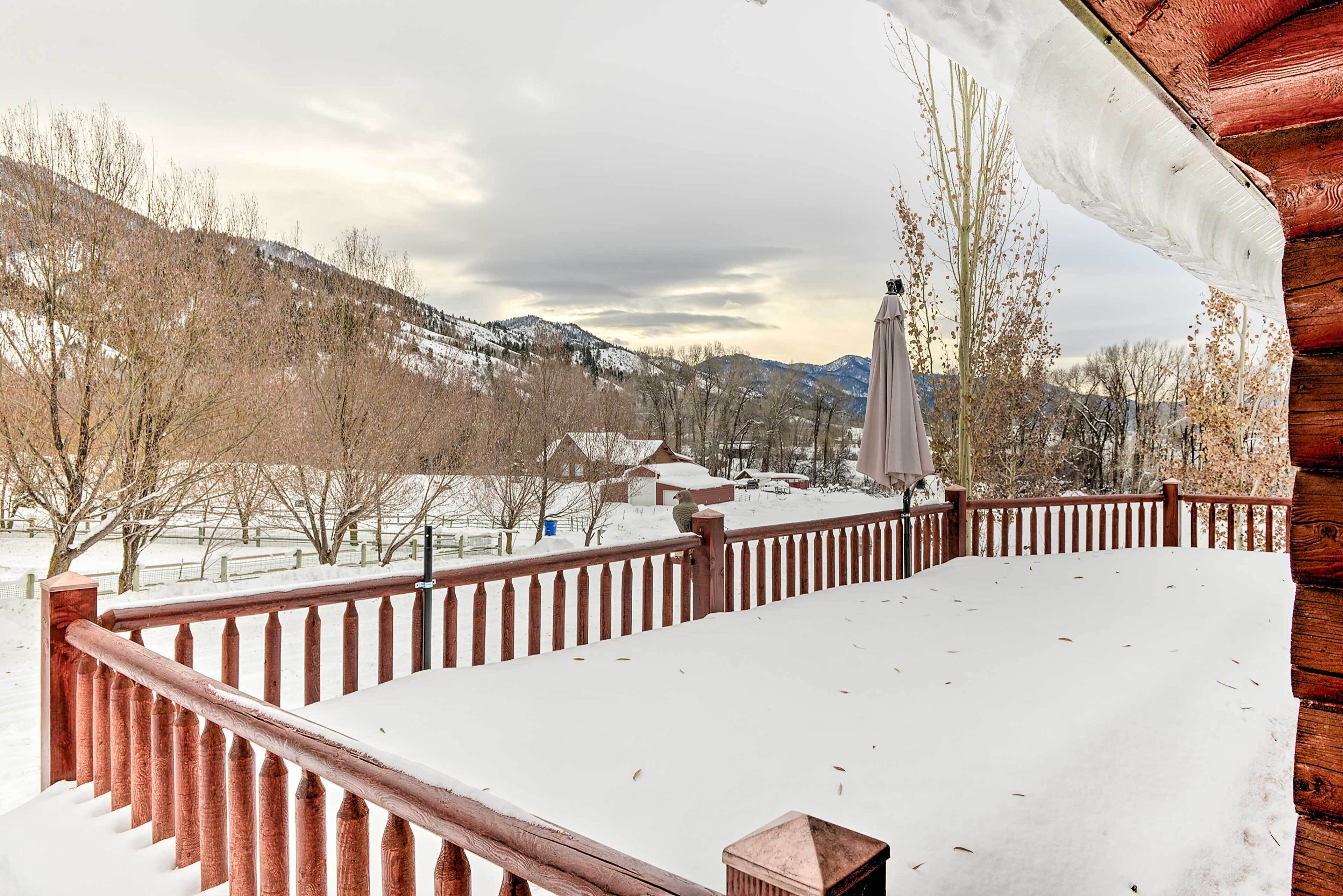 A wooden deck with a railing, snow-covered ground, and a view of a snowy landscape with trees and houses in the background.