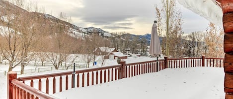 A wooden deck with a railing, snow-covered ground, and a view of a snowy landscape with trees and houses in the background.