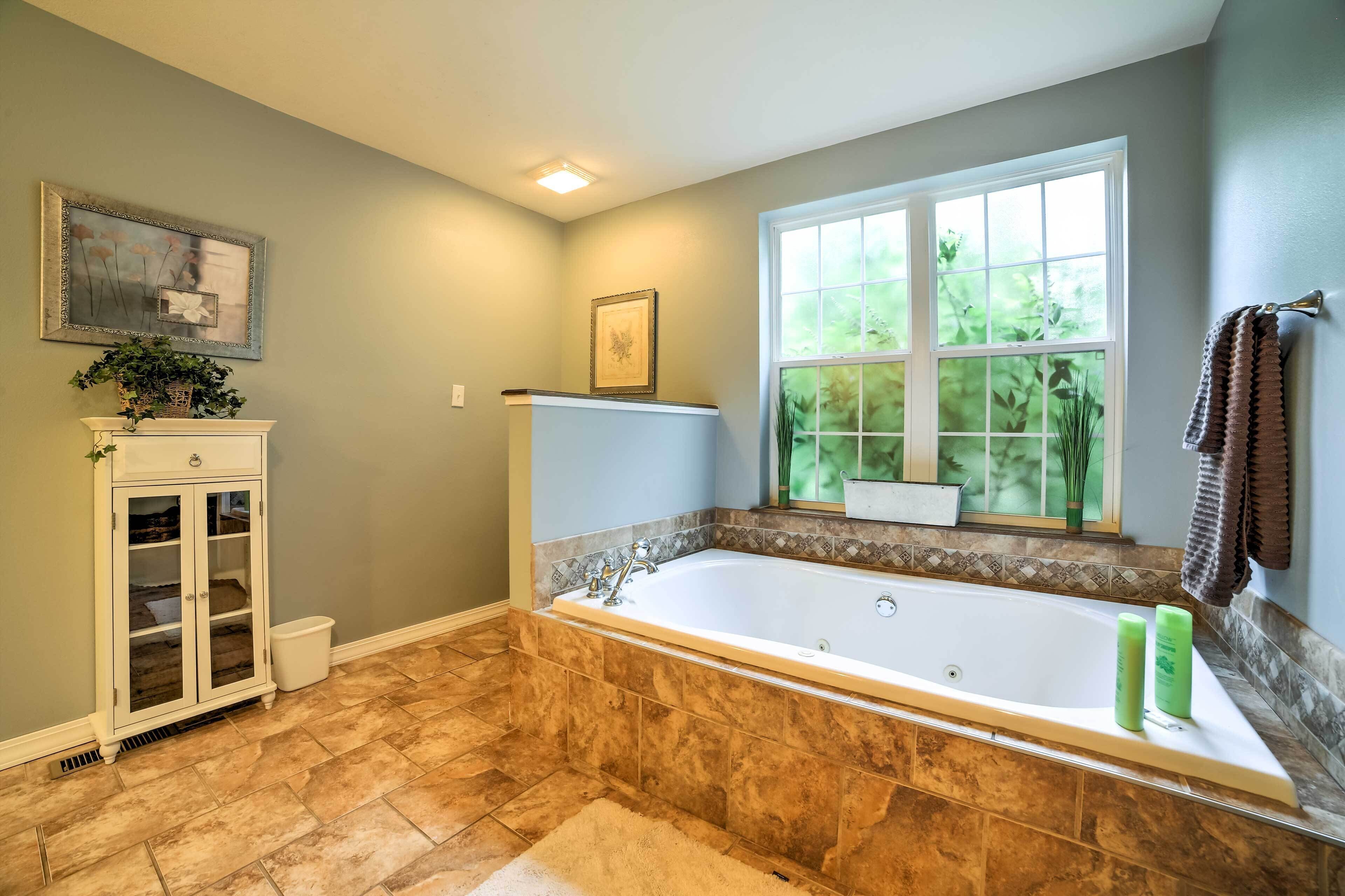 A bathroom with a large bathtub, a window with a view of greenery, a cabinet with glass doors, and a small plant on the cabinet.