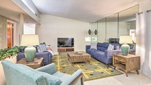 A living room with two sofas, a coffee table, and a TV. There is a large mirror on one wall and a window providing natural light. - Retro St Pete Home w/ Private Heated Pool (St. Petersburg)
