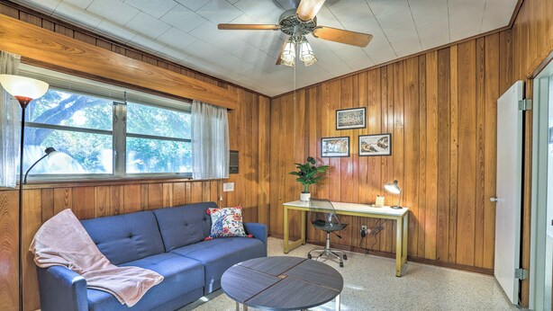A room with a wooden paneled wall, a blue sofa with a blanket, a small round table, a desk with a lamp, and a ceiling fan. - Retro St Pete Home w/ Private Heated Pool (St. Petersburg)