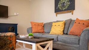 A living room with a grey sofa, orange and yellow cushions, a small table with a plant, and a wall-mounted TV. - Single-story Manchester Home Close to Parks! (Manchester)