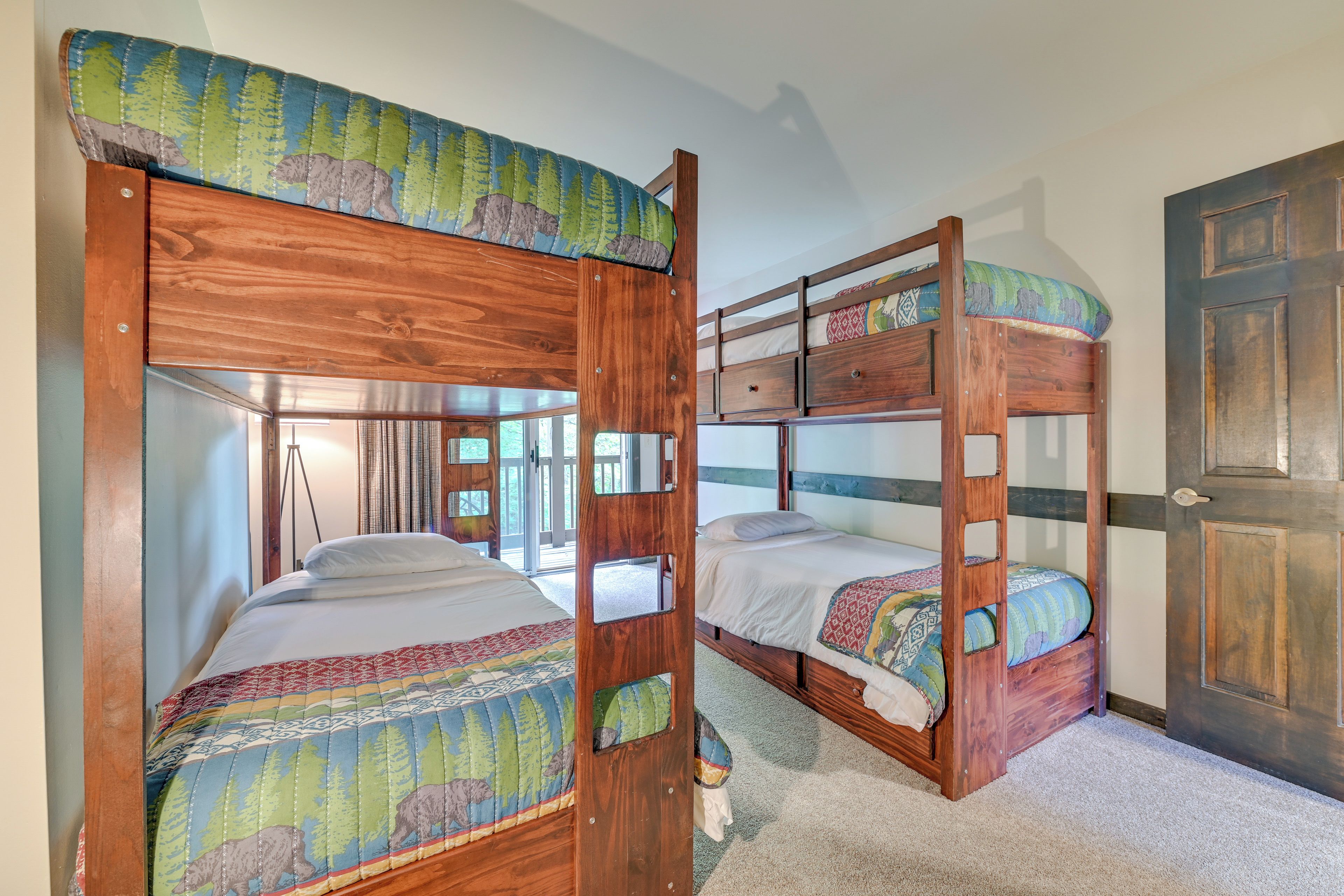 Une chambre avec deux lits superposés, chacun recouvert d’une couverture à motifs et doté d’une tête de lit ornée d’un décor forestier. On y trouve une porte en bois et une fenêtre avec des rideaux.