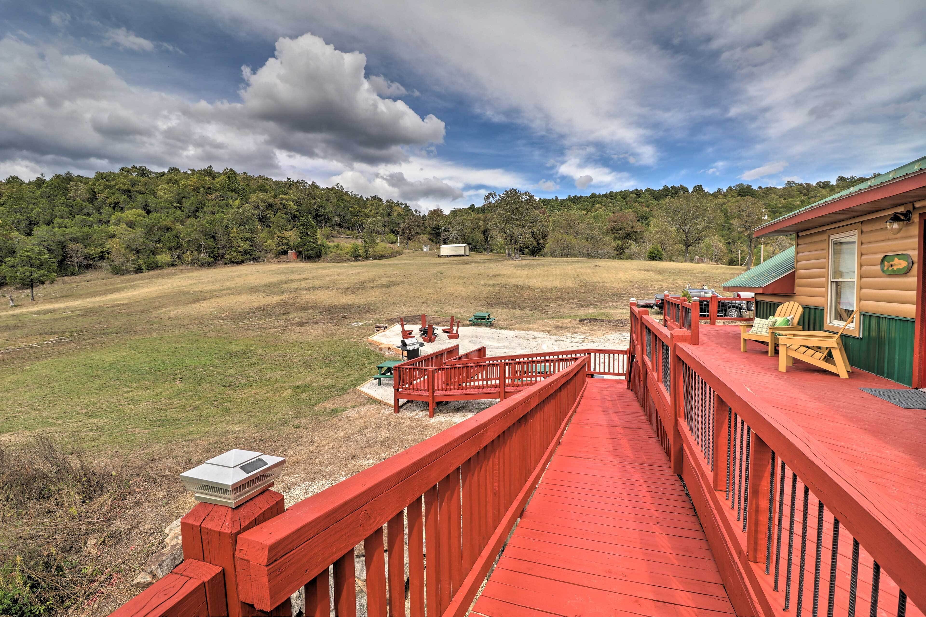 A wooden deck with a red railing overlooking a grassy field and a cabin with a green roof.
