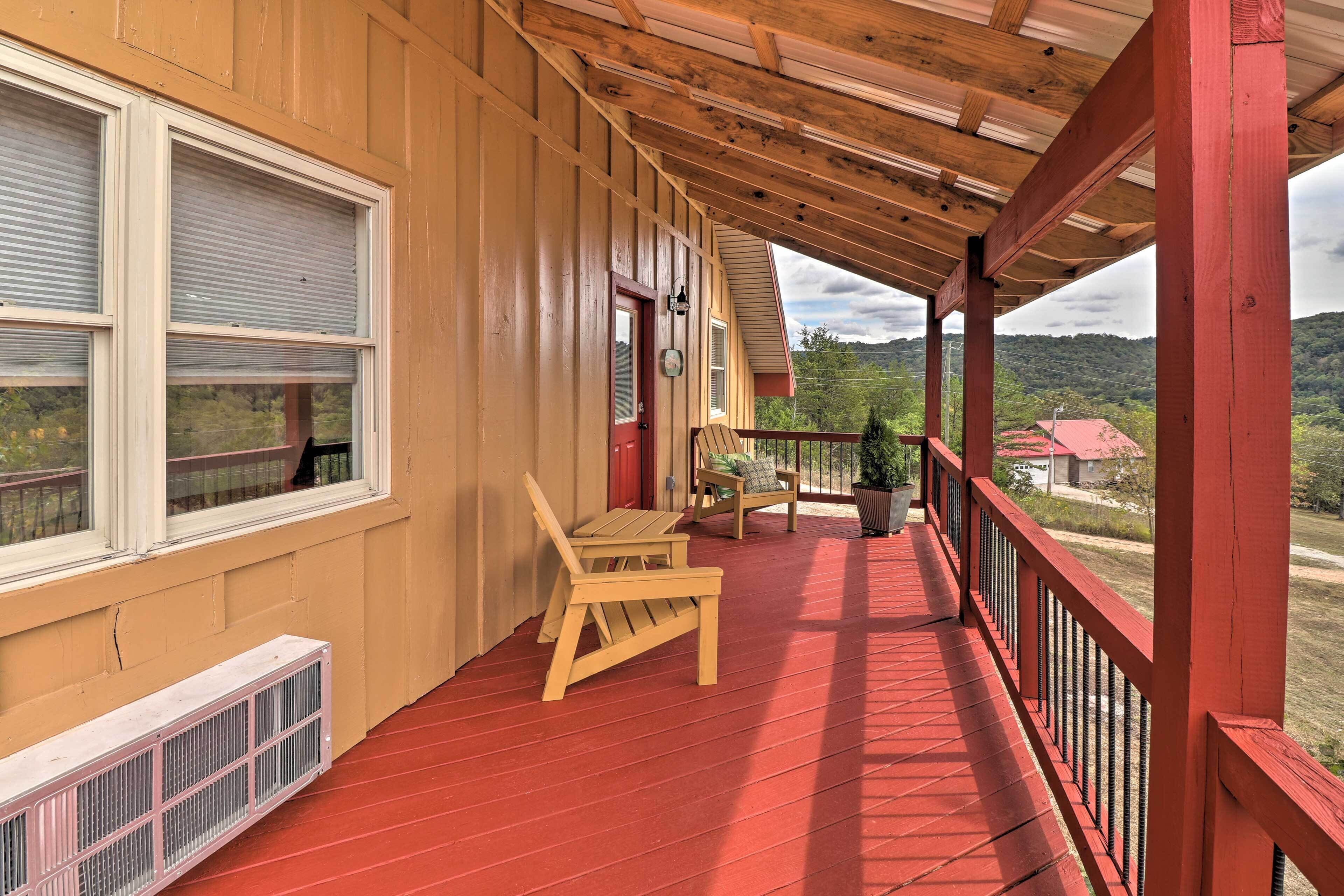 A covered porch with wooden chairs and a red railing, overlooking a green landscape.