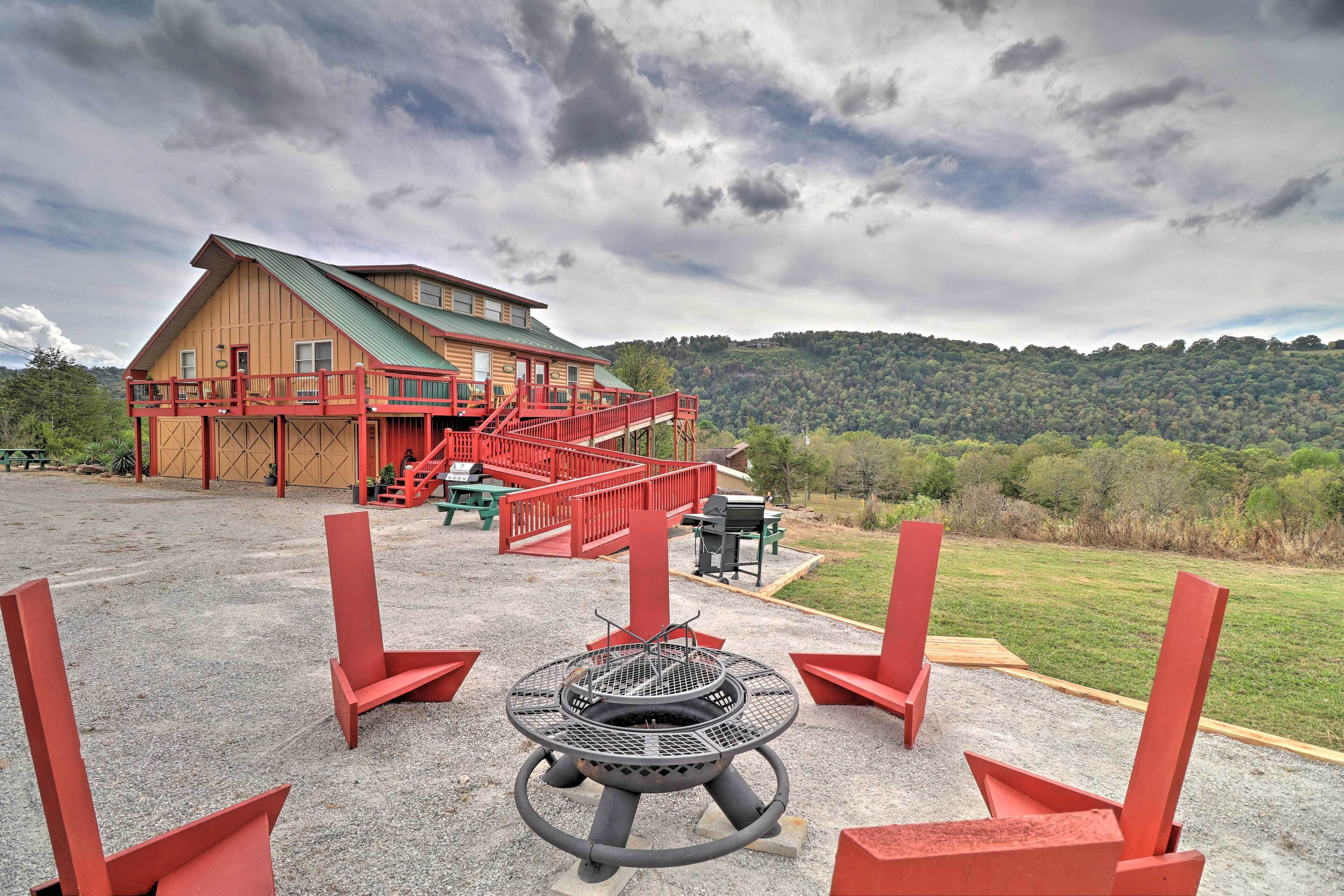 Un chalet doté d’un foyer extérieur et de chaises rouges devant, entouré d’un paysage pittoresque composé de collines et d’un ciel nuageux.