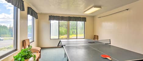 A room with a ping pong table, a chair, a plant, and large windows.