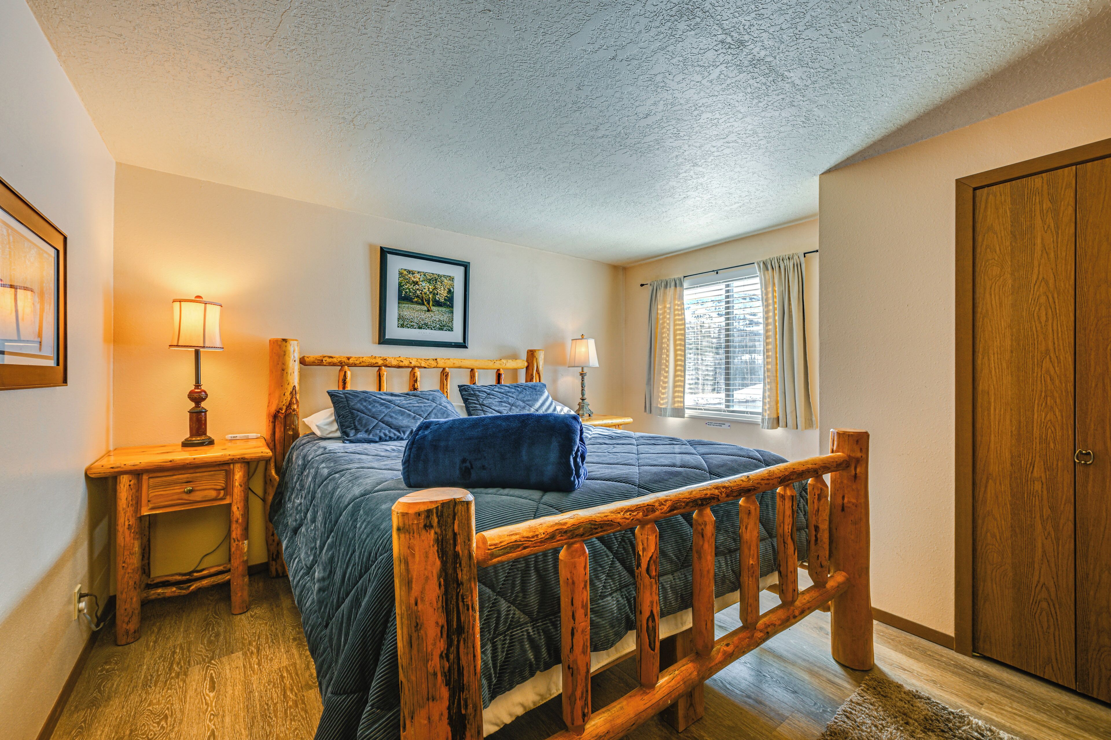 A bedroom with a wooden bed frame, a bedside table with a lamp, a window with curtains, and a door.
