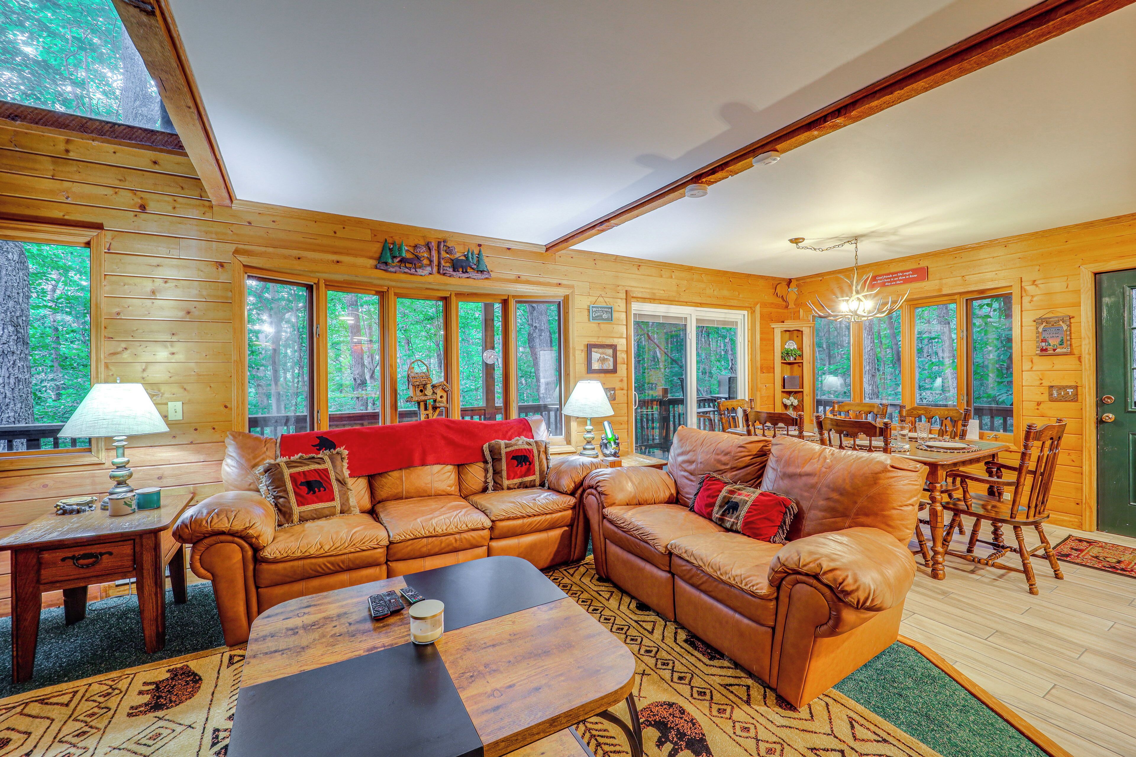 A cozy cabin interior with wooden walls, large windows, a dining area, and a living room with leather sofas and a coffee table.