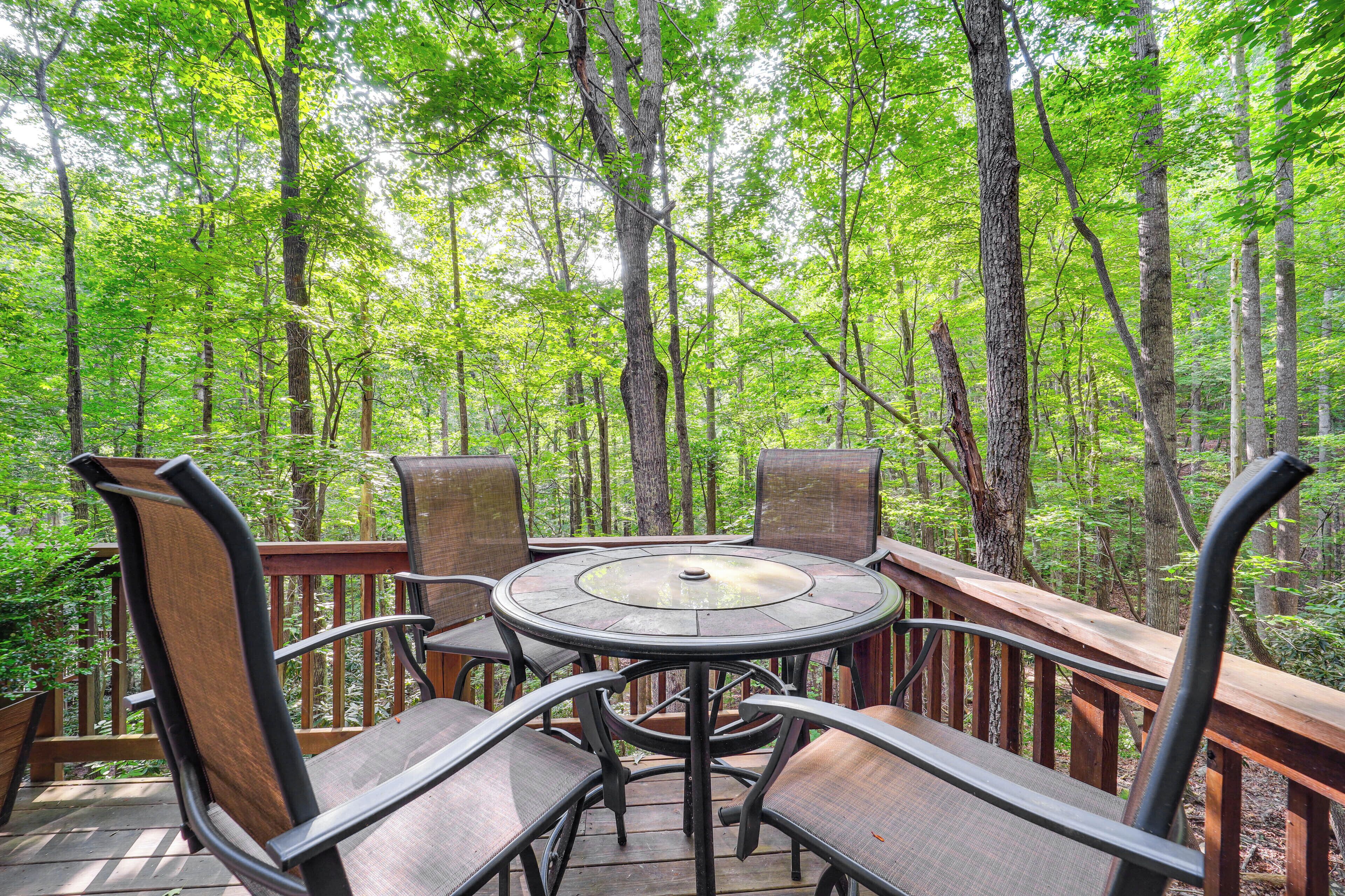 A round wooden table with metal legs surrounded by cushioned chairs on a deck with a wooden railing, set in a forested area.