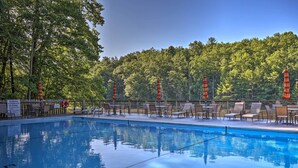 Piscina com superfície de água azul cristalina, rodeada por espreguiçadeiras e mesas, com um cenário de vegetação densa ao fundo.