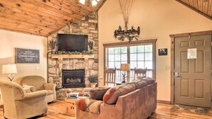 A cozy living room with a stone fireplace, wooden ceiling, and a large window. - Screened Deck: Cozy Branson West Cabin! (Branson West)