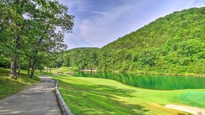 A paved pathway alongside a green golf course with a lake and forested hills in the background. - Screened Deck: Cozy Branson West Cabin! (Branson West)