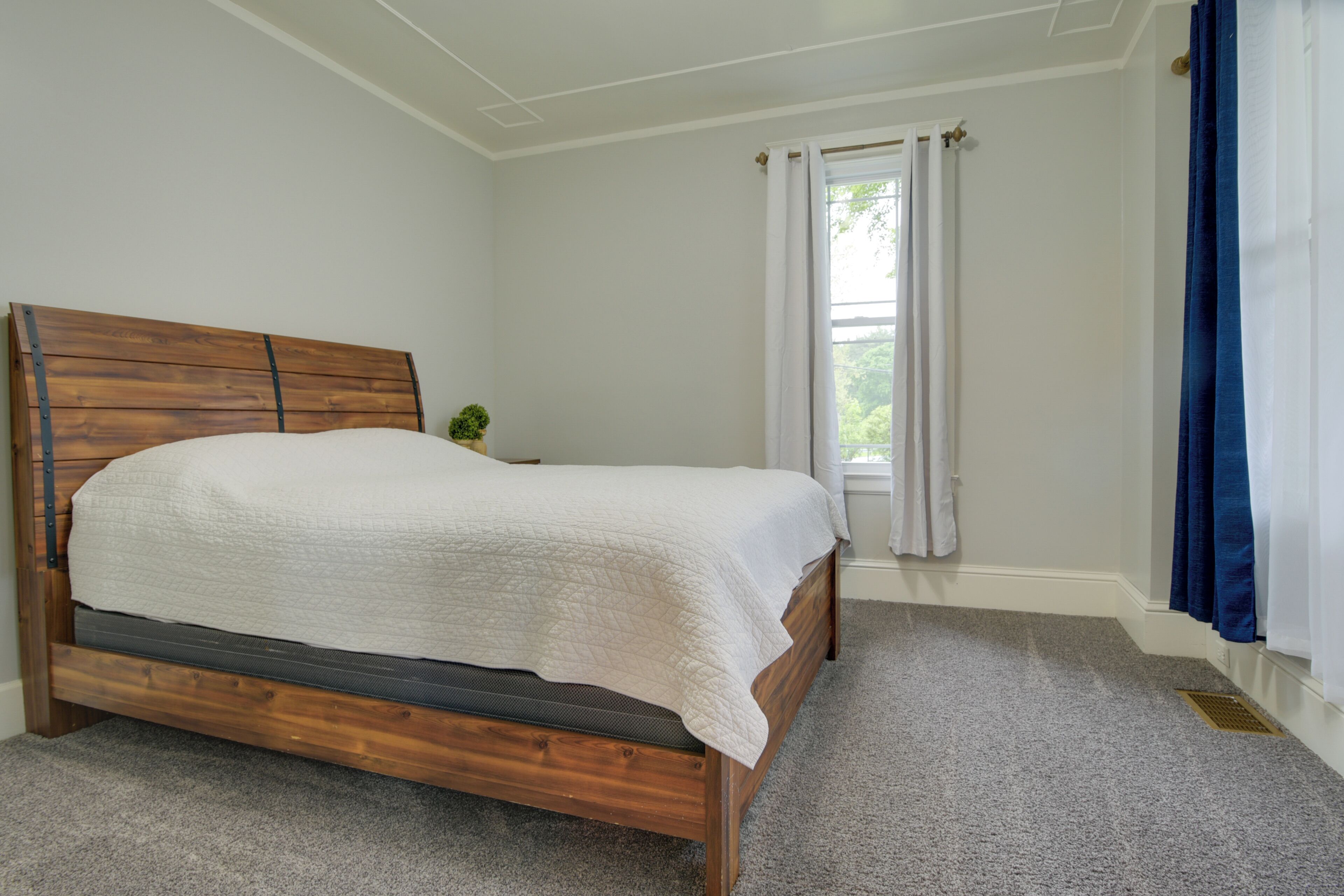 A bedroom with a wooden bed, white walls, a window with white and blue curtains, and a small plant on a nightstand.