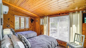 Habitación de cabaña de madera con cama, dos ventanas, una silla y aire acondicionado instalado en la pared.