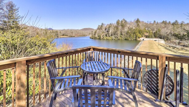 Una terraza de madera con mesa y sillas con vista a un lago, rodeada de árboles y colinas.