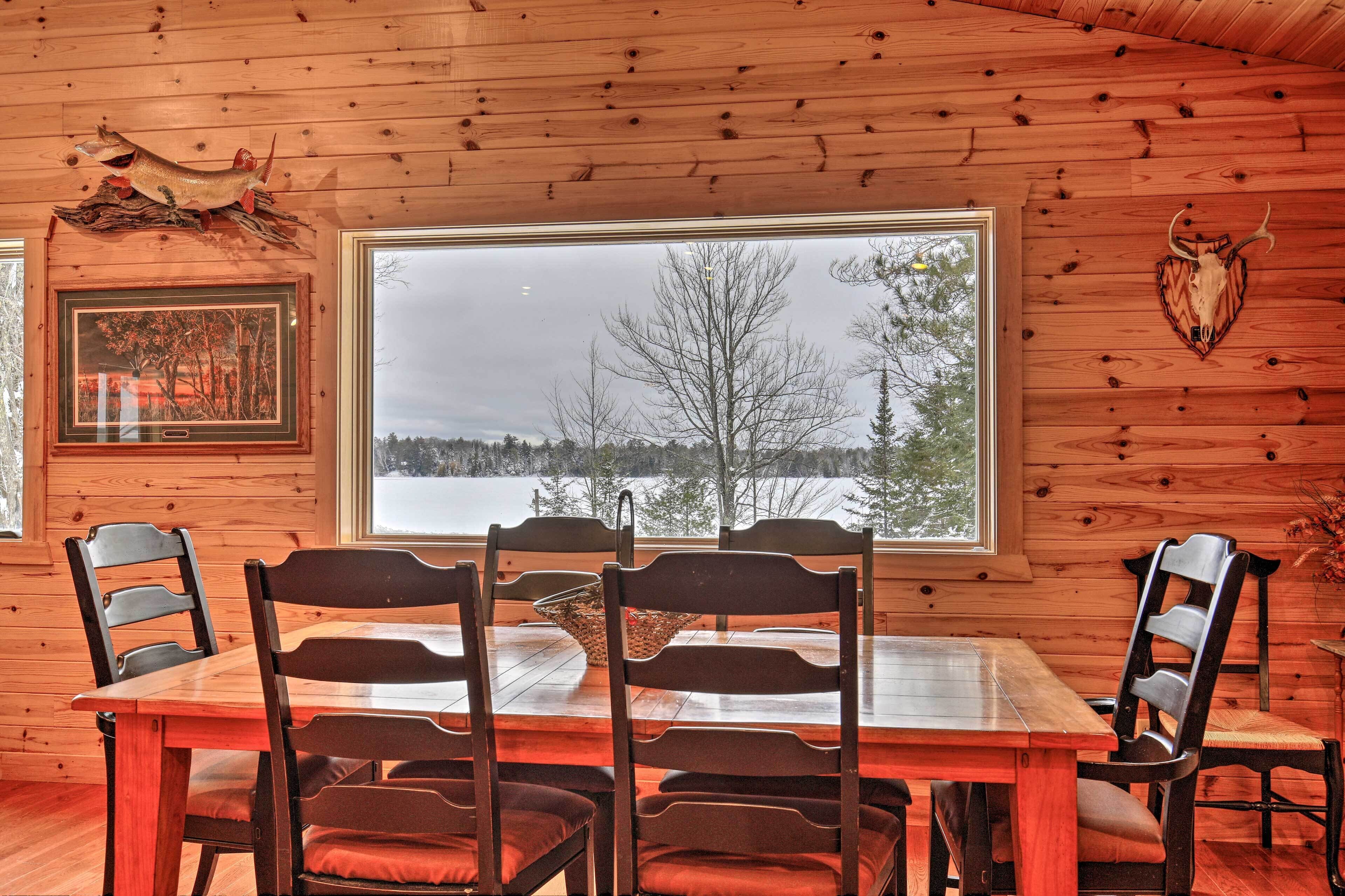 A wooden cabin interior with a dining table and chairs, a large window with a view of trees and a snowy landscape, and a mounted animal skull on the wall.