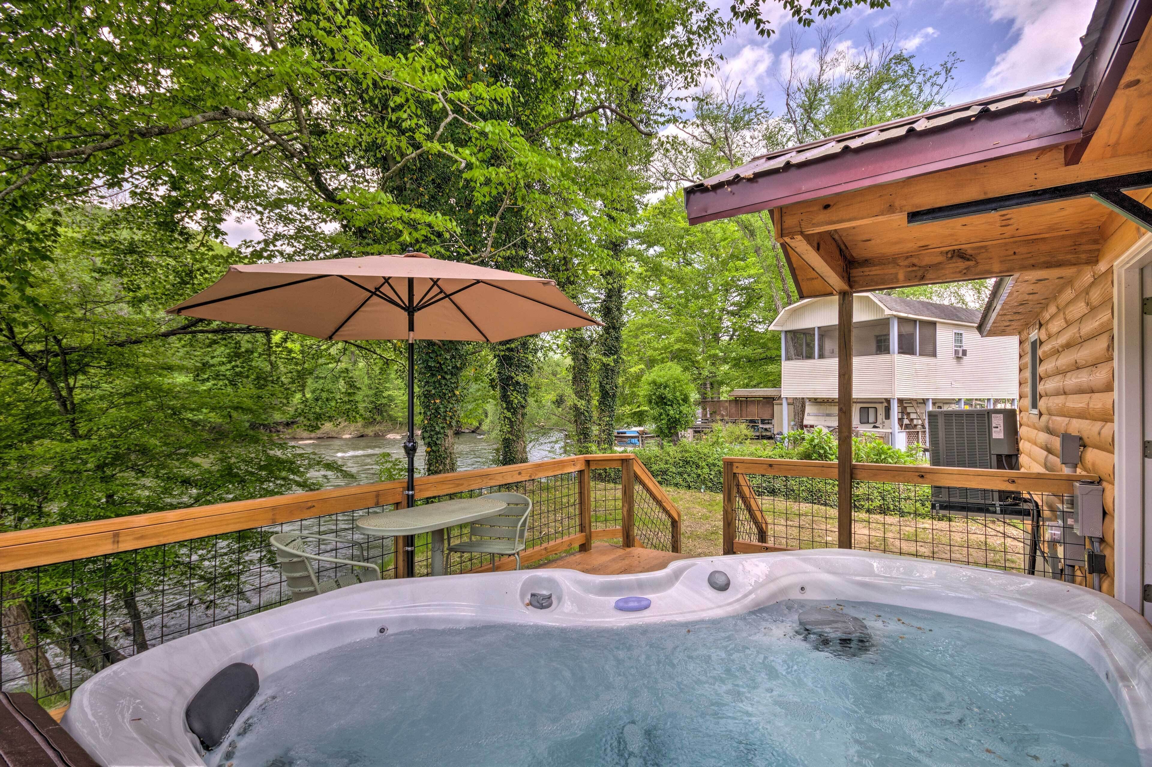 Un jacuzzi en una terraza de madera con una mesa y una sombrilla, con vista a un río y una casa.