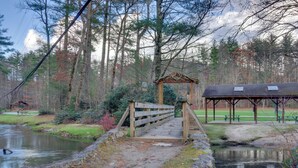 Un puente de madera sobre un estanque, con un área de picnic y un refugio al fondo.