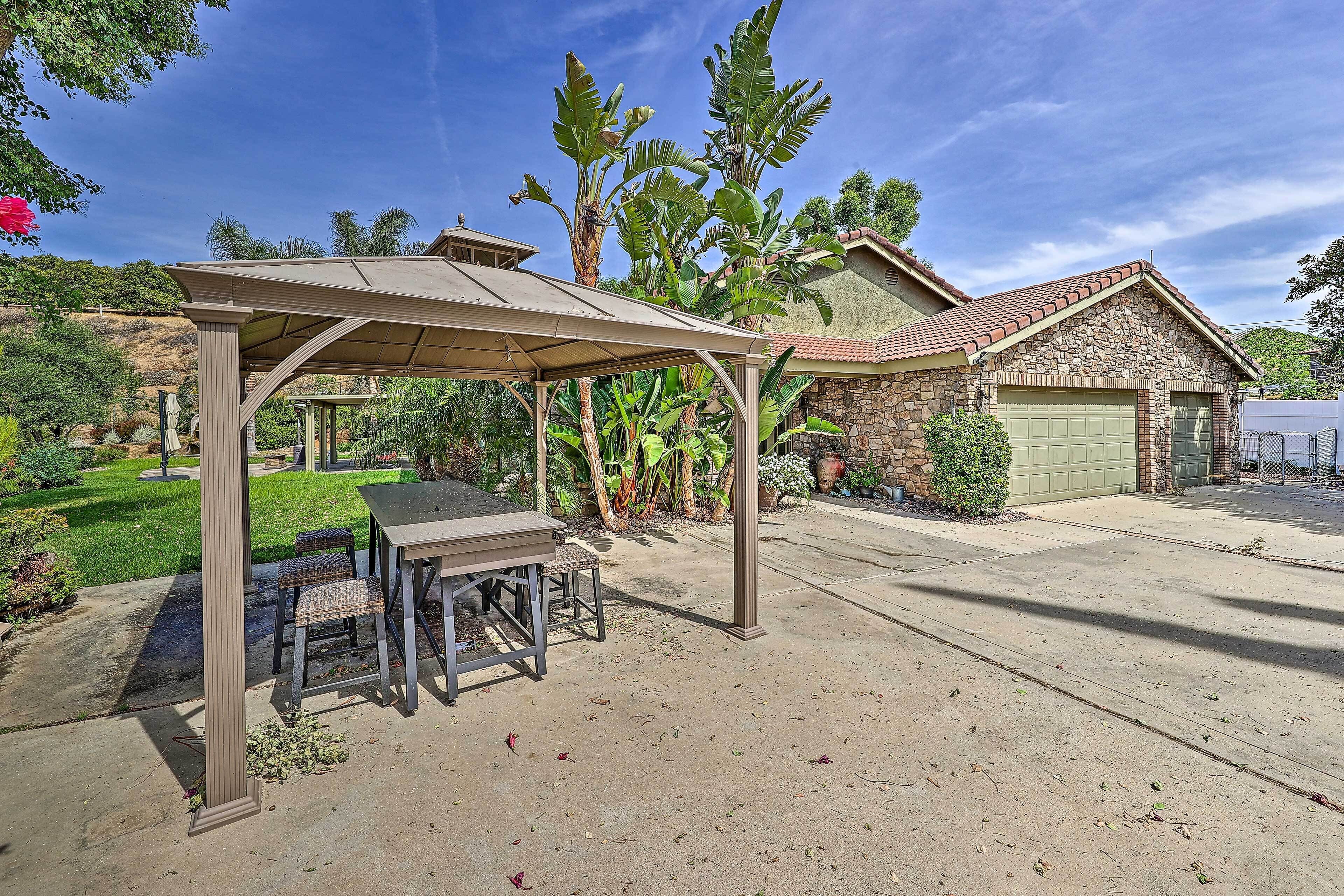 Eine überdachte Terrasse mit Tisch und Stühlen, ein Haus aus Stein mit Garage und üppiges Grün im Hintergrund.