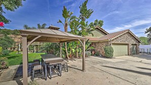 A covered patio with a table and chairs, a stone house with a garage, and lush greenery in the background. - Spacious & Private Wine Country Villa w/ View (Temecula)