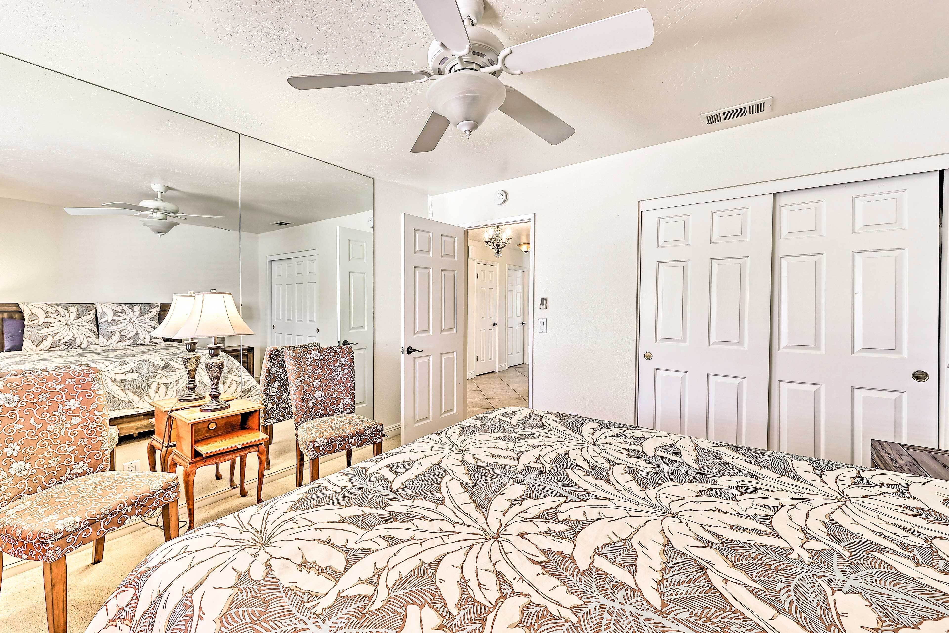 A bedroom with a bed, a ceiling fan, a desk with a lamp, a chair, and a large mirror.